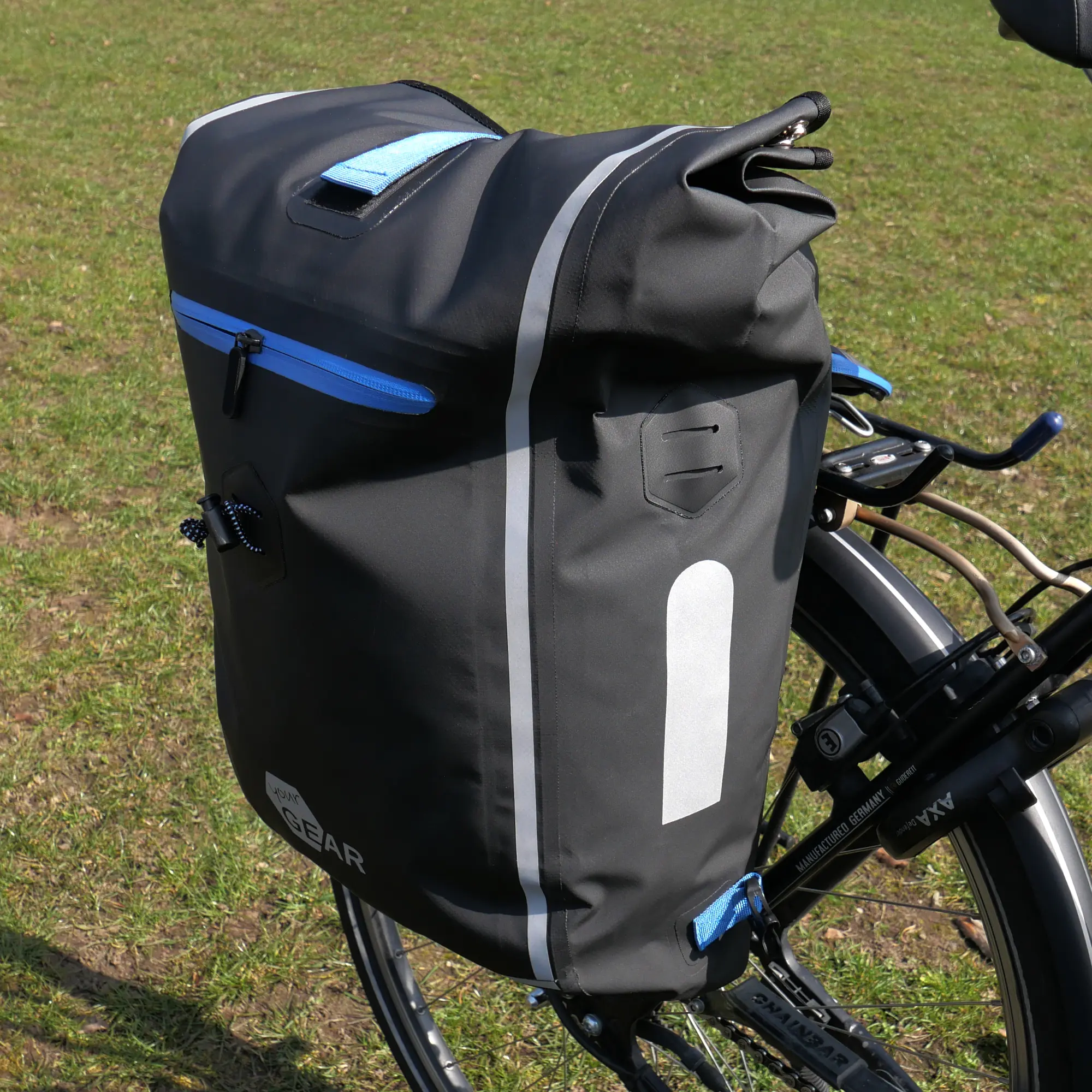 yourGEAR Arco 19 L waterproof 3-in-1 bike pannier, shown on a bicycle rack. Black bag with blue zipper, reflective stripes, roll-top closure, and yourGEAR logo, suitable as backpack or laptop office bag.