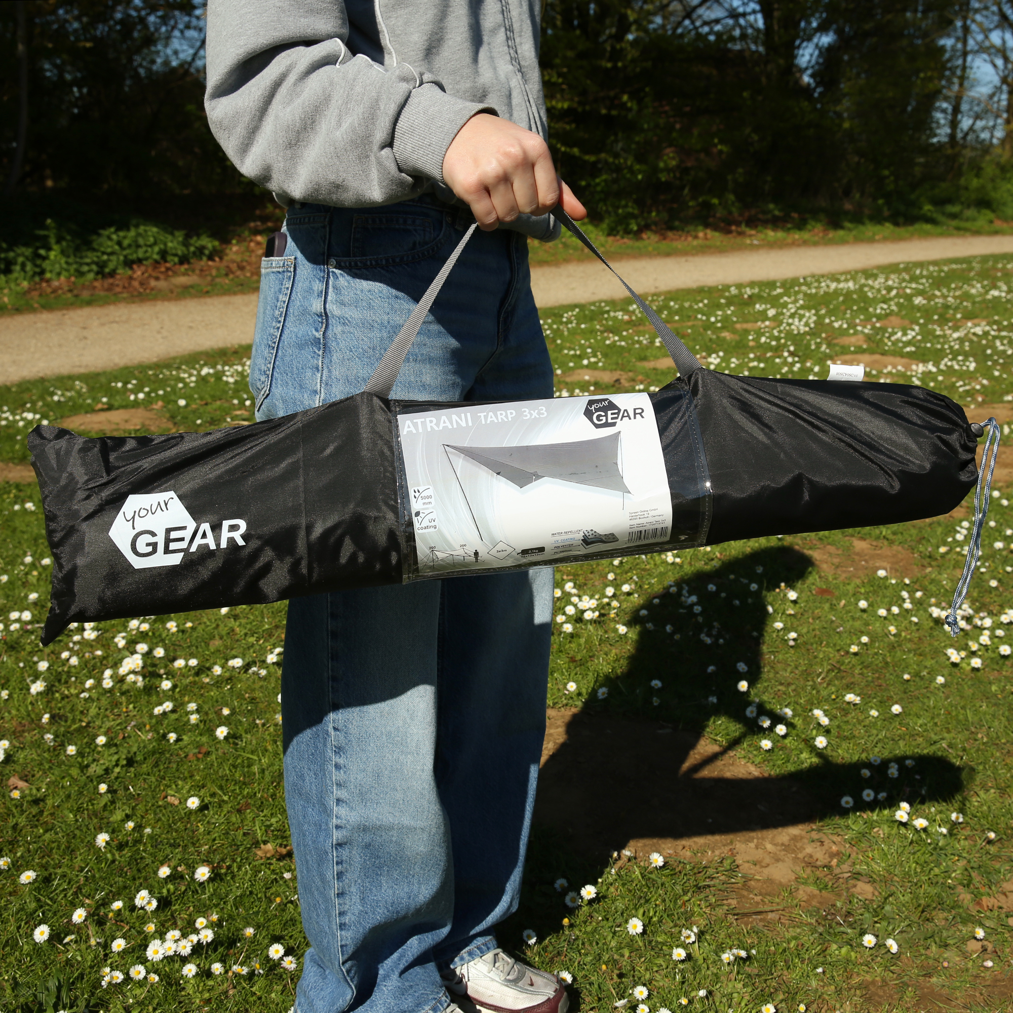 Persona che porta la sacca nera del Tarp your GEAR "Atrani 3x3", etichetta visibile, maniglia grigia, borsa da trasporto per telo parasole impermeabile grigio, accessori inclusi, scena all'aperto su prato.