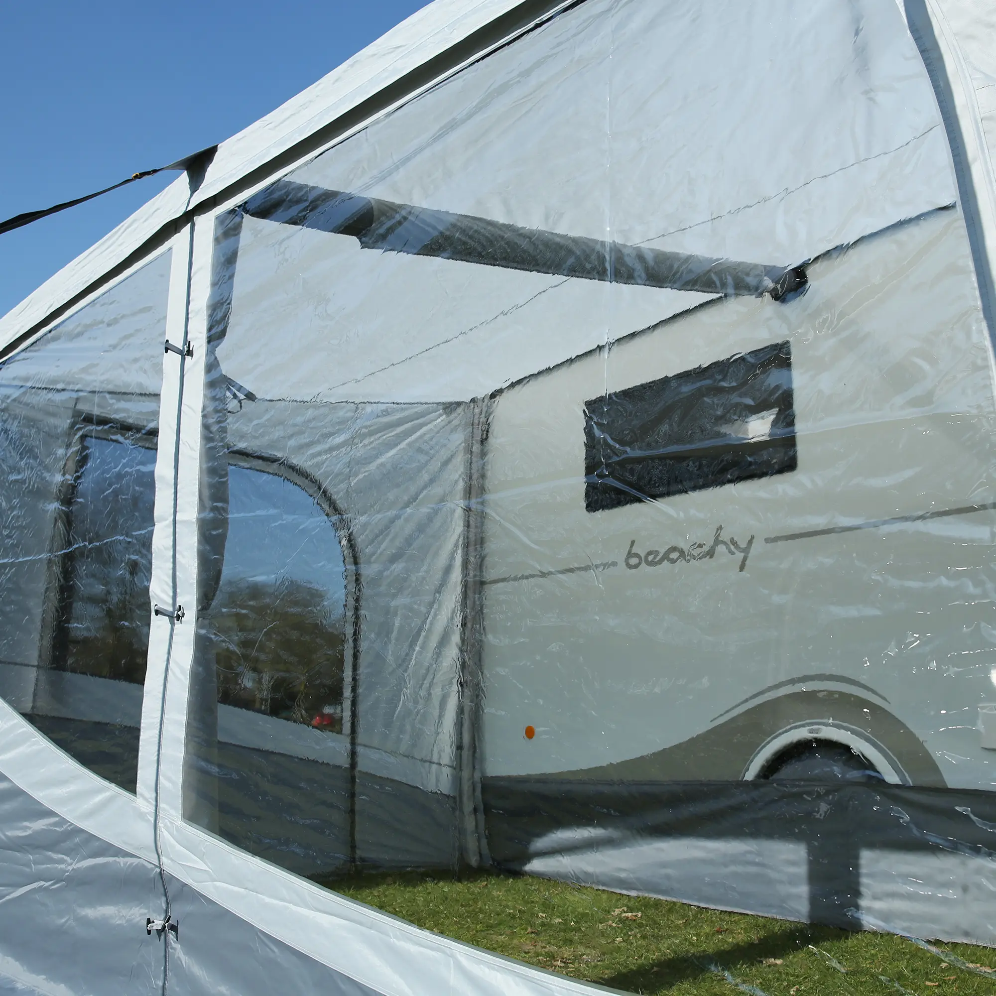 Vue rapprochée d'une caravane blanche derrière la paroi transparente d'une tente ou d'un auvent sous un ciel bleu.