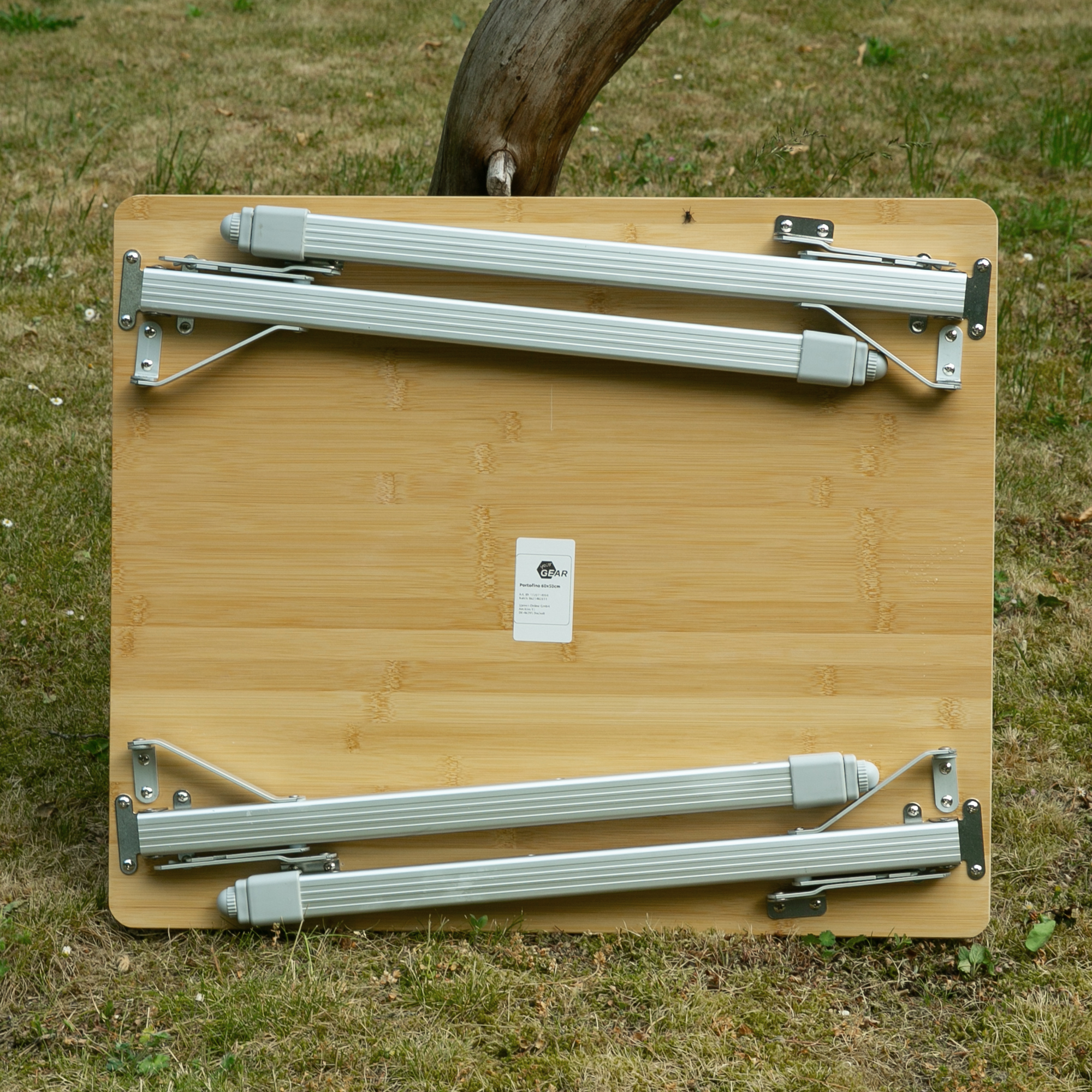 Bambus Campingtisch your GEAR Portofino M mit klappbaren Aluminium-Teleskopbeinen, zusammengeklappt auf Rasen stehend; robuste, glatte Bambusplatte, sichtbare Scharniere und Sicherungen, rechteckige Form.