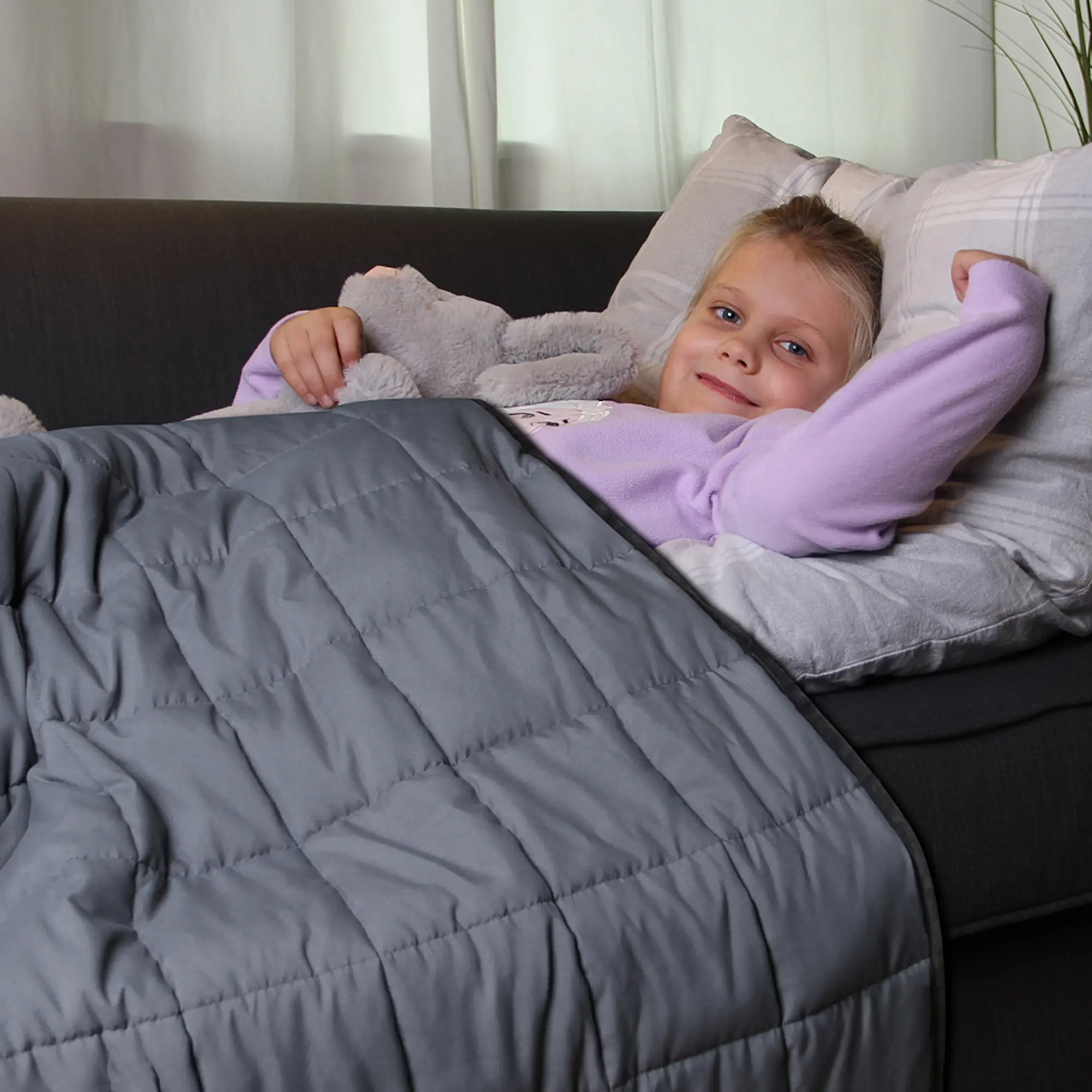 Niña acostada en un sofá, cubierta con la yourGEAR Como Therapiedecke, manta de peso infantil de 3 kg y 100% algodón, color gris, tamaño 100x150 cm, ideal para relajación y terapia de presión profunda.