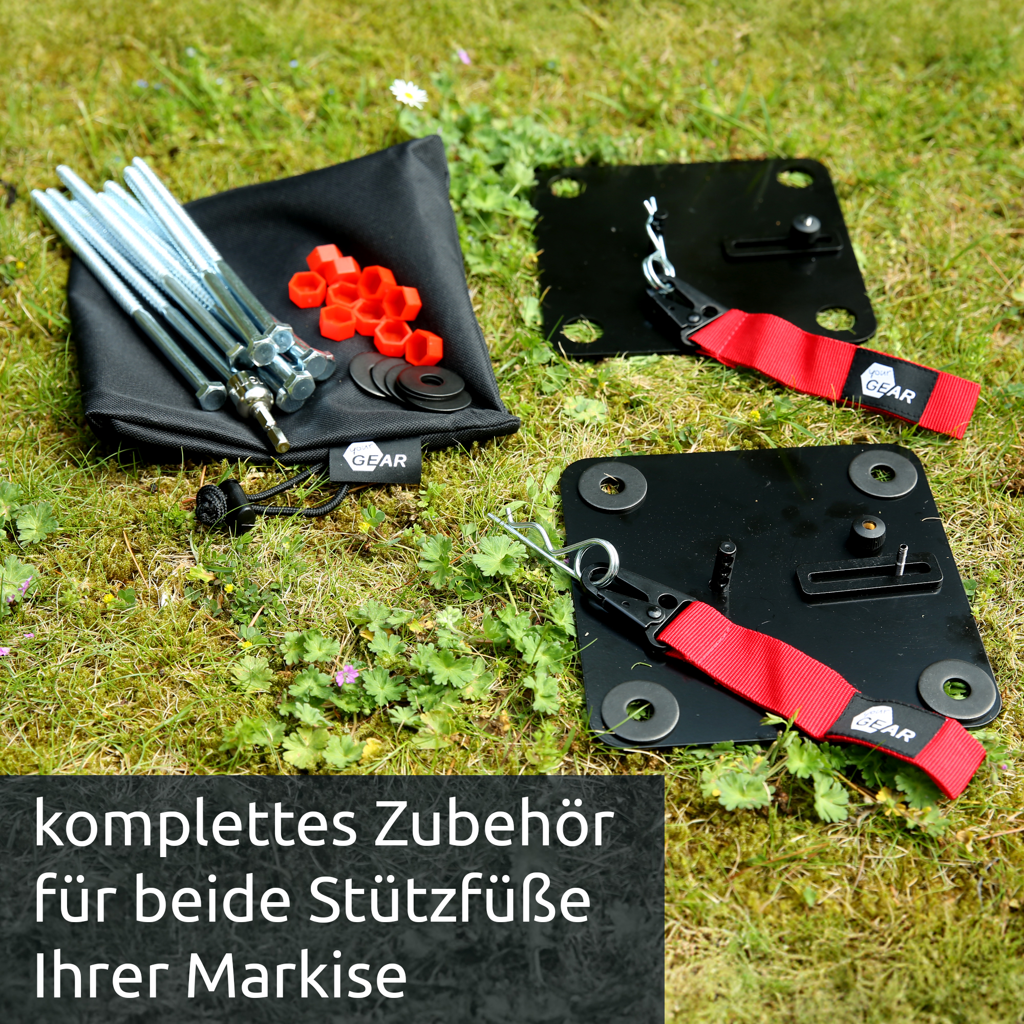 Black steel anchor plates with red webbing strap, zinc-coated long bolts, orange plastic nut caps, black washers and small hardware laid on grass — your GEAR Montana awning ground anchor kit for camper/caravan.