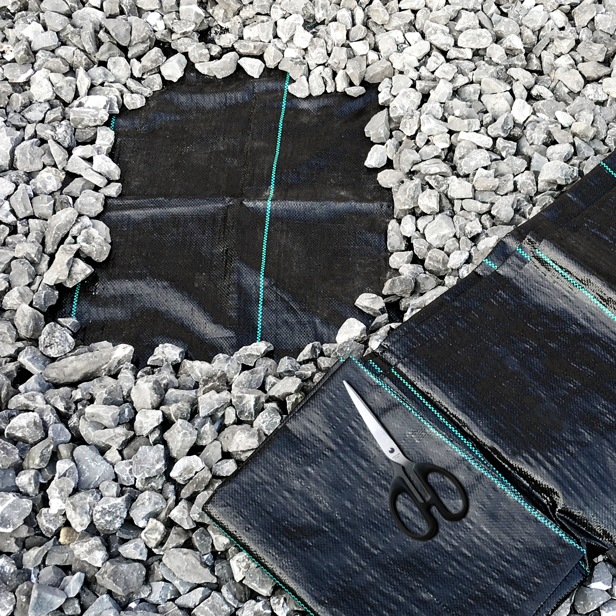 yourGEAR weed control fabric, 10 m², shown as black, tear-resistant garden fleece with green stripes, laid under gravel. Scissors placed on the fabric, highlighting easy cutting and installation.
