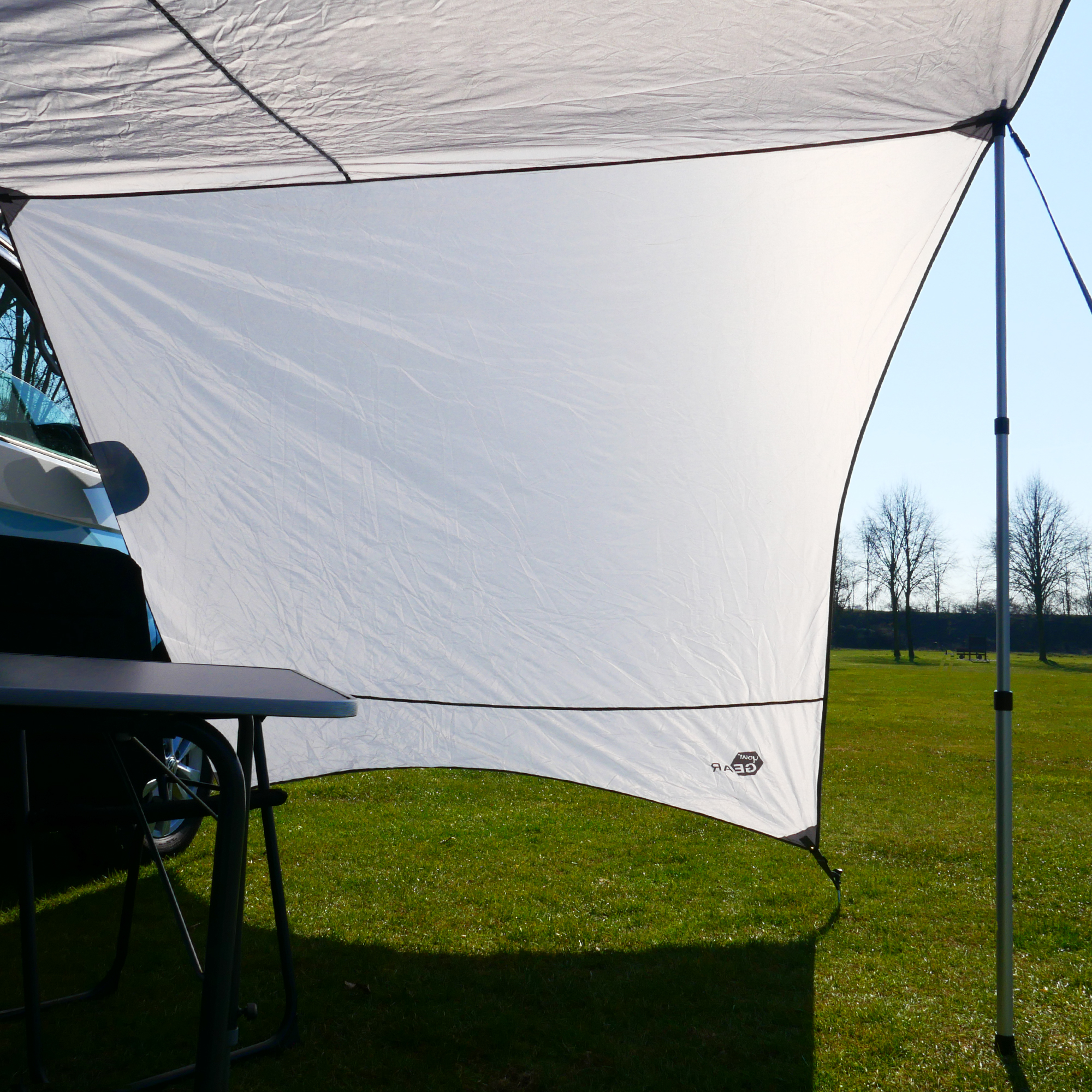 your GEAR Tropea Bus awning UV 50+ tarp with sidewalls, shown mounted on a van. Cool gray fabric, aluminum telescopic poles, providing sun and rain protection for camping or outdoor use. Table and chair underneath.