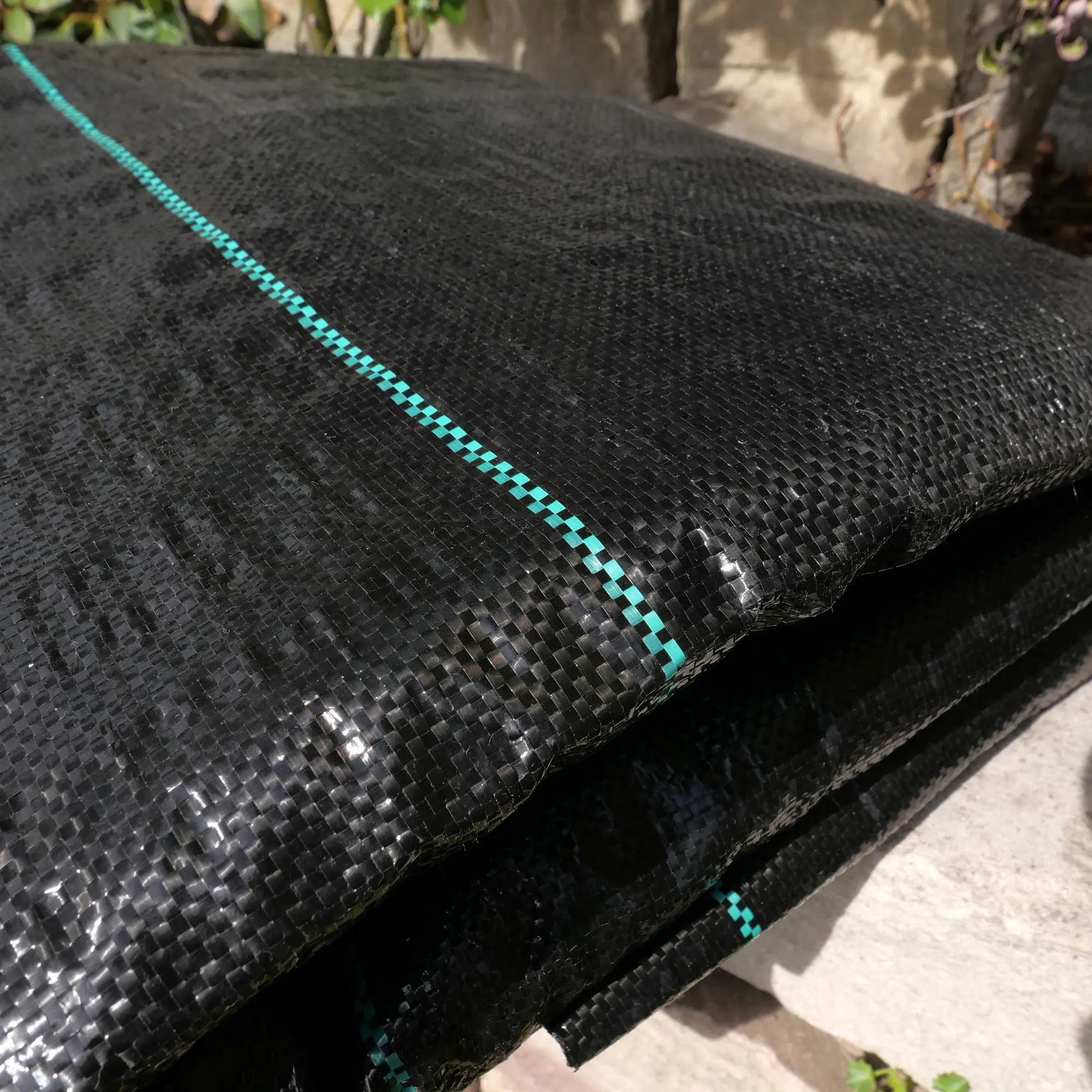 Toile de paillage anti-mauvaises herbes yourGEAR 10 m² noire, tissée, avec ligne verte, résistante, perméable et stable aux UV, format 5x2 m, pliée et posée sur le sol de jardin.