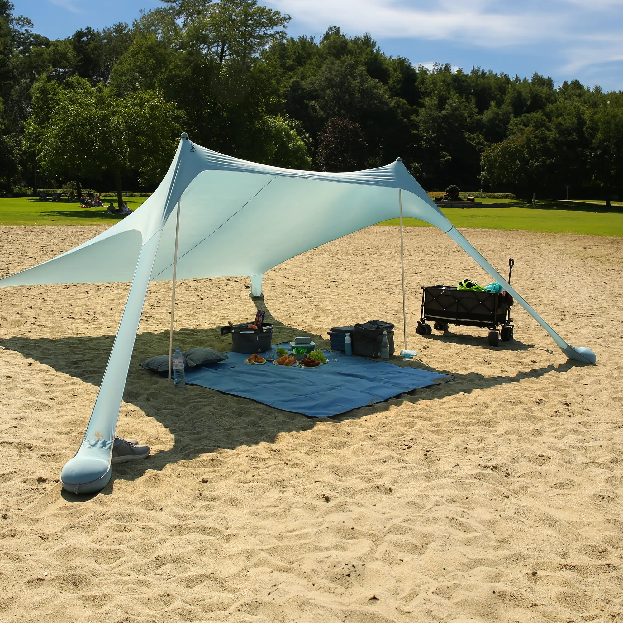 Toldo de playa your GEAR Cameo 3x3 m XXL en color azul claro, hecho de lycra elástica, montado en la arena con postes y cuerdas, ofrece amplia sombra y protección UV 50+ para picnic y descanso al aire libre.