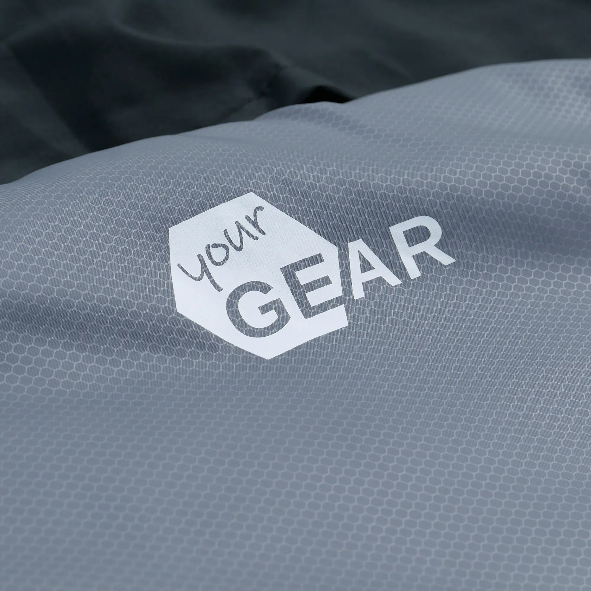 Primer plano del logo yourGEAR impreso en tela gris con patrón hexagonal, parte del saco de dormir doble Ostria Duo azul y gris, diseñado para dos personas y uso en verano.