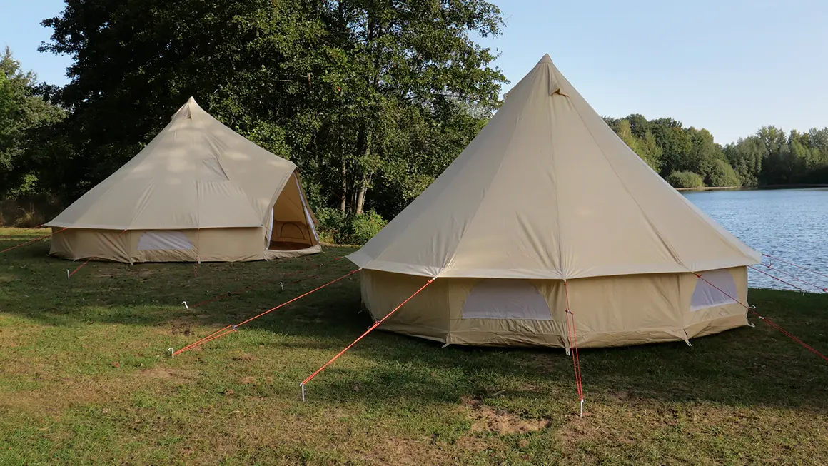 Zwei große beige Zelte stehen auf einer Wiese am Seeufer, umgeben von Bäumen und Natur.