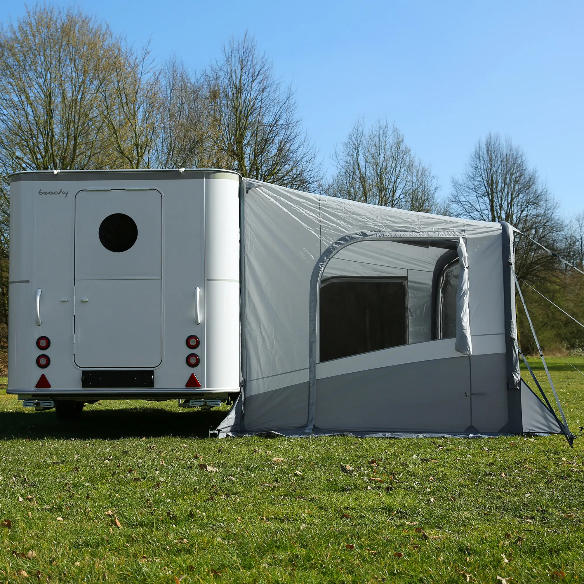 Caravane blanche avec une grande tente grise installée à l’arrière, garée sur une pelouse verte sous un ciel bleu, entourée d’arbres sans feuilles.