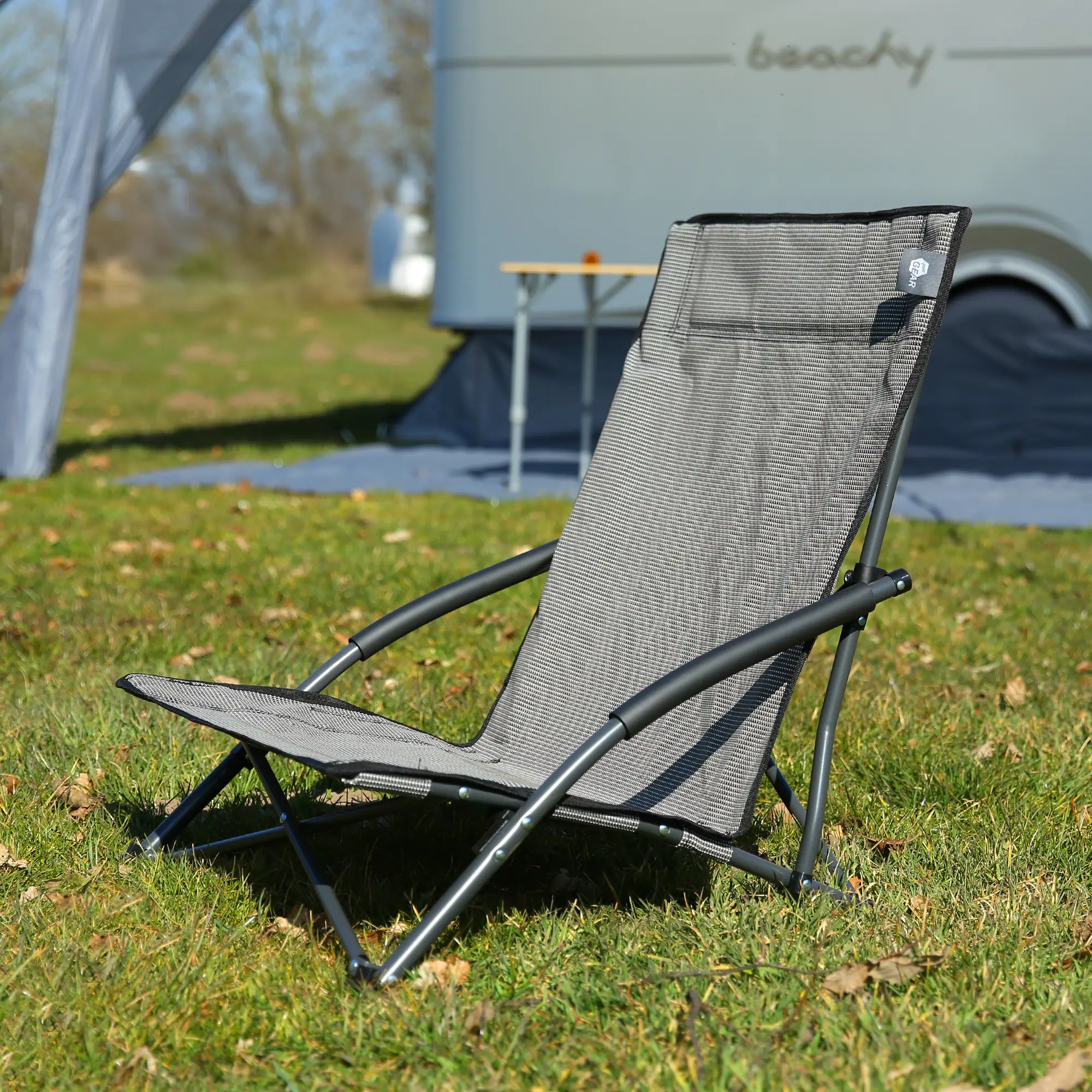 Silla de playa your GEAR Olbia de textileno gris con respaldo alto, marco metálico negro, reposacabezas acolchado y asiento bajo, ubicada sobre césped al aire libre, ideal para camping y jardín.