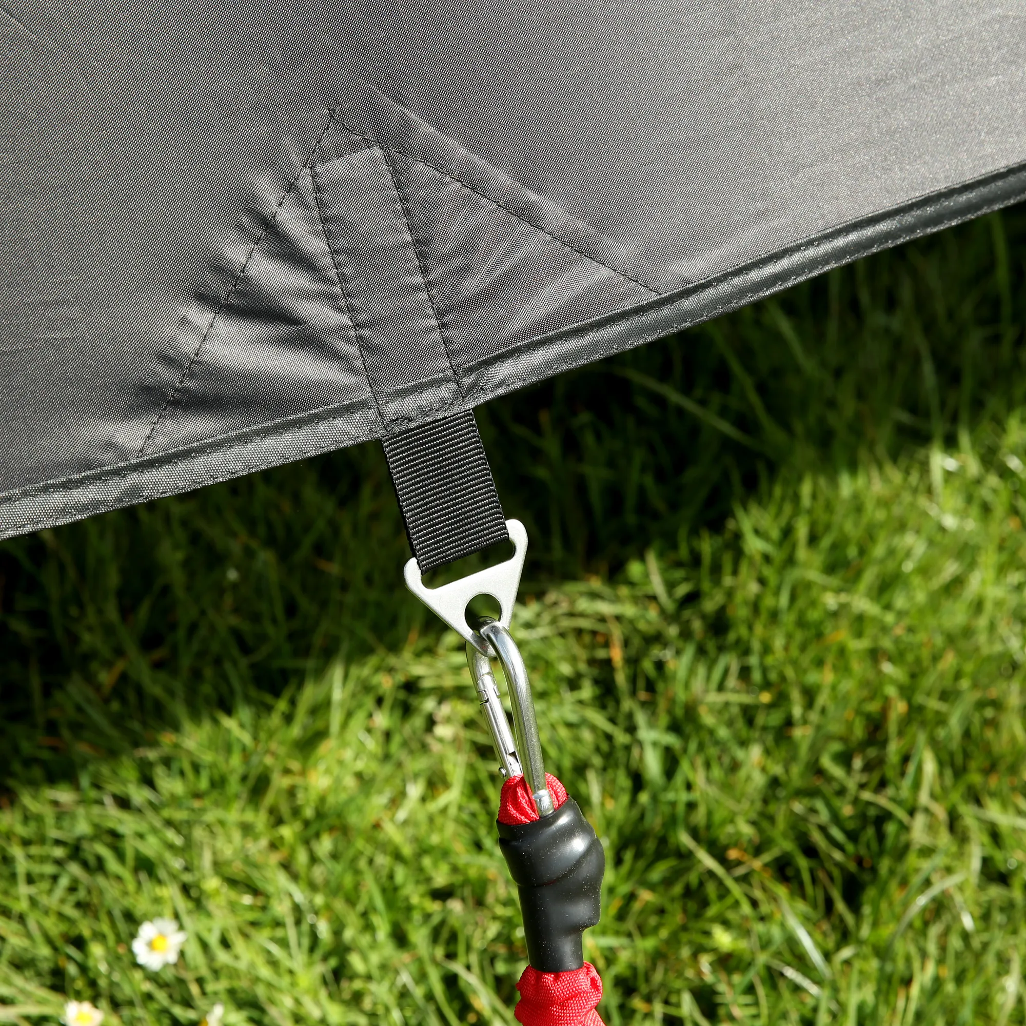 Close-up of grey your GEAR Phoenix awning front panel corner with reinforced stitching, black webbing strap and metal triangular anchor clip attached to a carabiner and red bungee cord over green grass