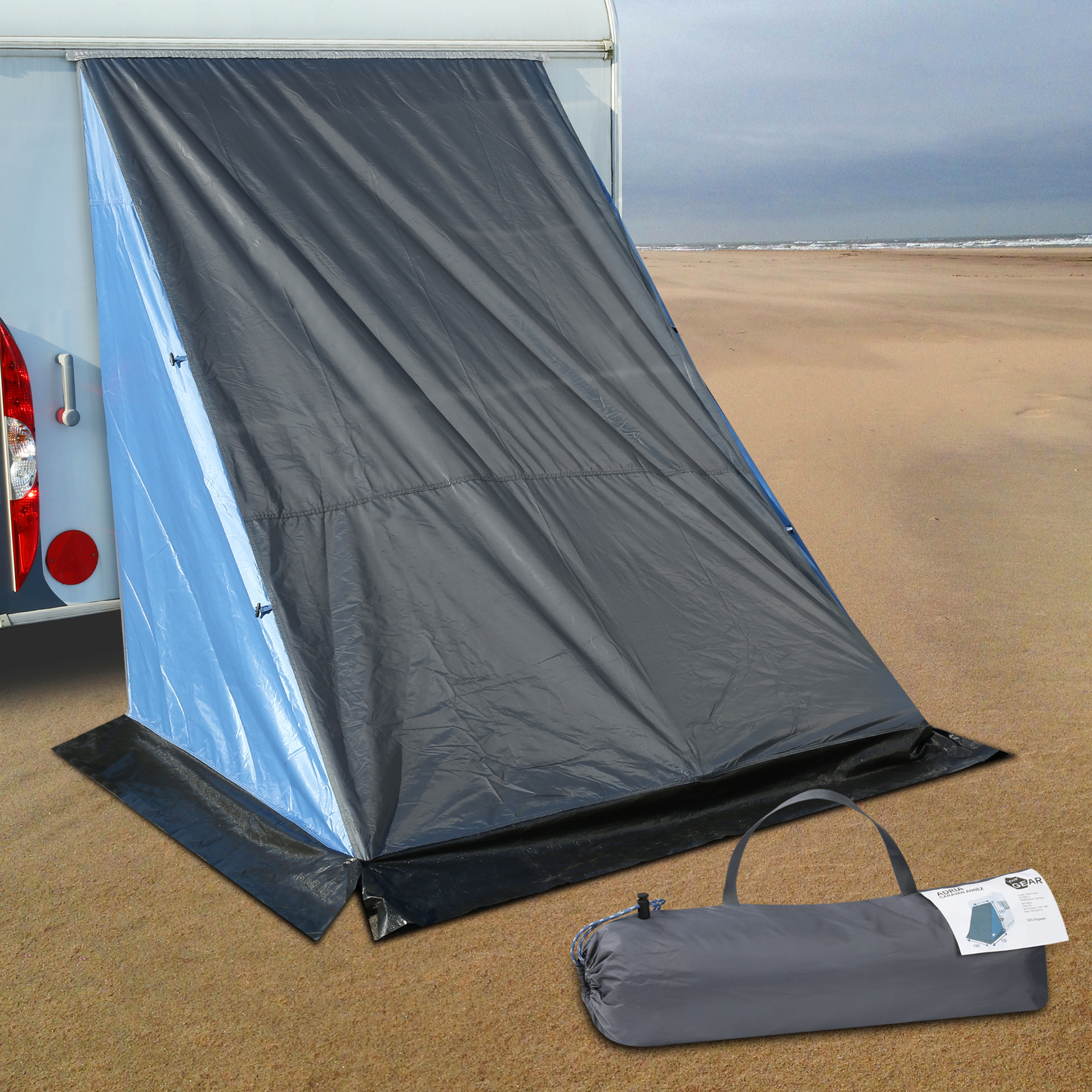 Tente de hayon your GEAR Adria 180 fixée à l’arrière d’une caravane, offrant un espace de stockage protégé. Tissu gris, bordure noire au sol, sac de transport inclus au premier plan sur le sable.