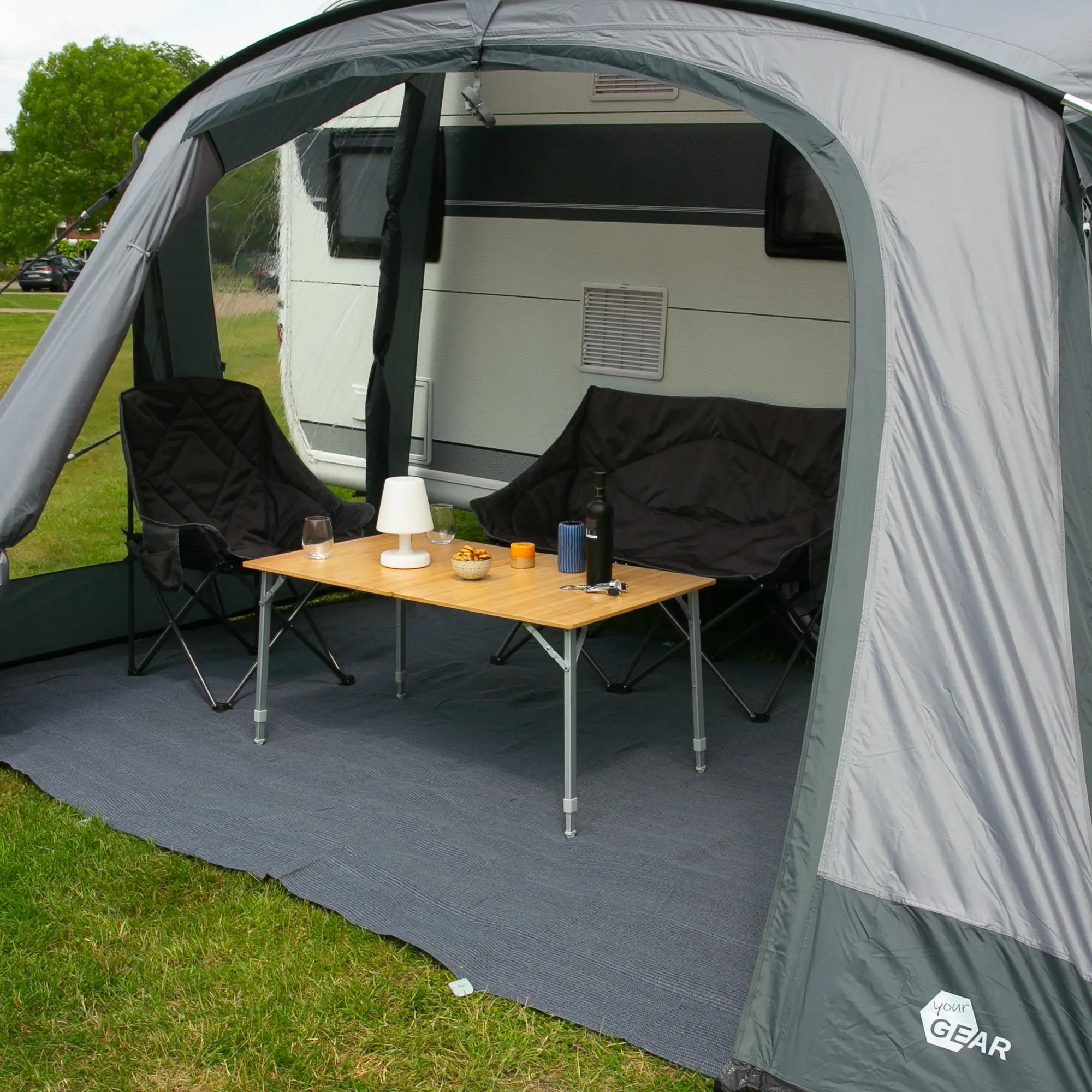 Bambus Campingtisch your GEAR Portofino XL für 6 Personen, rechteckig, höhenverstellbare Teleskopbeine, stabile Konstruktion, aufgestellt im Vorzelt vor einem Wohnwagen, mit Campingstühlen und Accessoires.