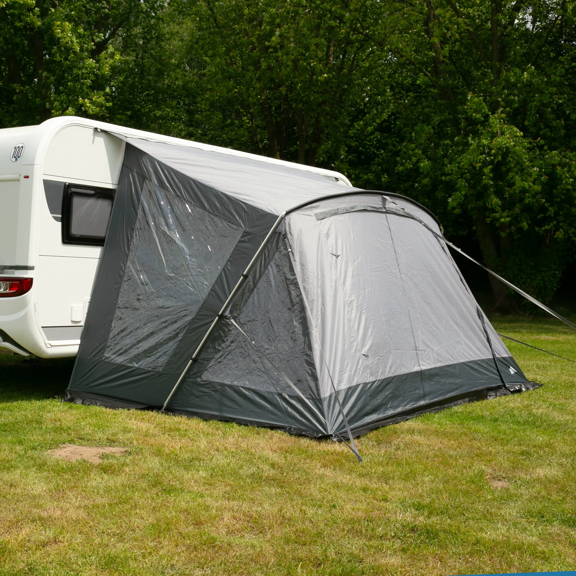 Großes your GEAR Vorzelt Positano 400 am Wohnwagen, mit vier Fenstern, großer Tür, stabilem Aluminium-Gestänge und UV-Schutz 50+, auf einer Wiese vor grünem Baumbestand aufgebaut.