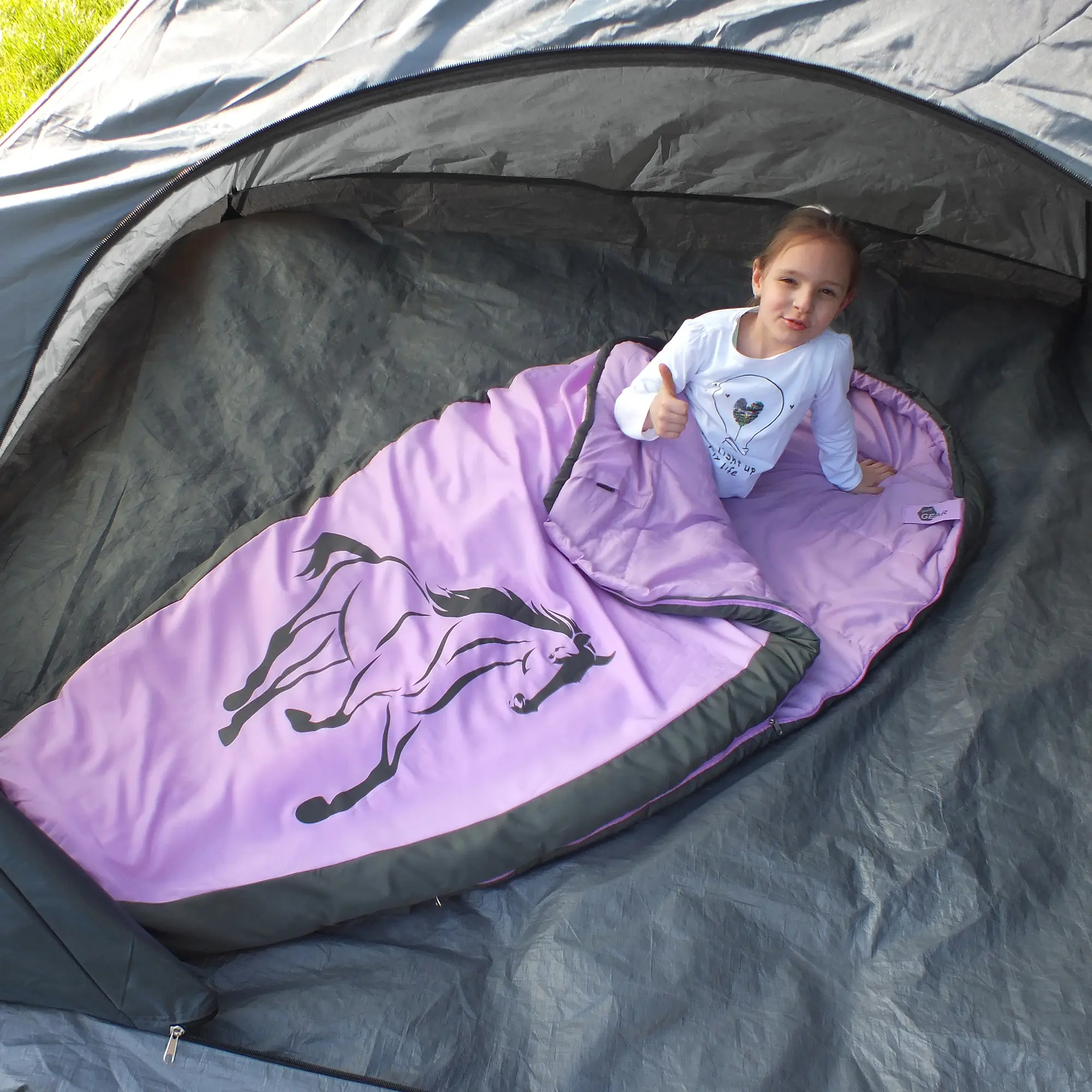 Fille dans un sac de couchage yourGEAR Horse, forme momie, rose et gris, motif cheval, intérieur doux, posé dans une tente. Sac adapté aux enfants, 180x90 cm, idéal pour l’été, rembourrage 300 g/m².