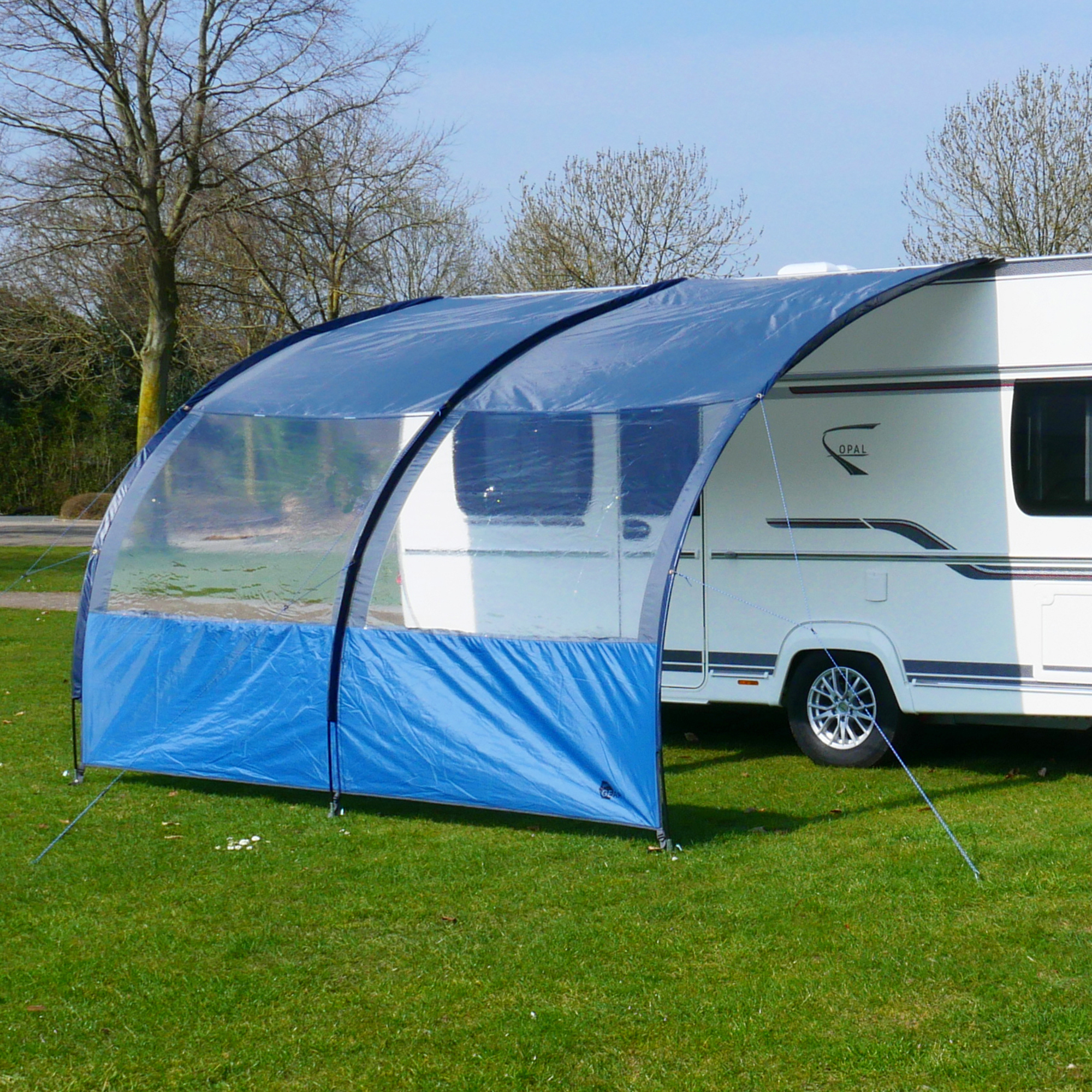 Vorzelt your GEAR Riviera 350, 3,5x2,6m, mit UV 50+ Schutz, großem Panoramafenster und blauem Wetterschutzdach, aufgebaut an einem Wohnwagen auf einer Wiese, ideal für Camping und Outdoor.