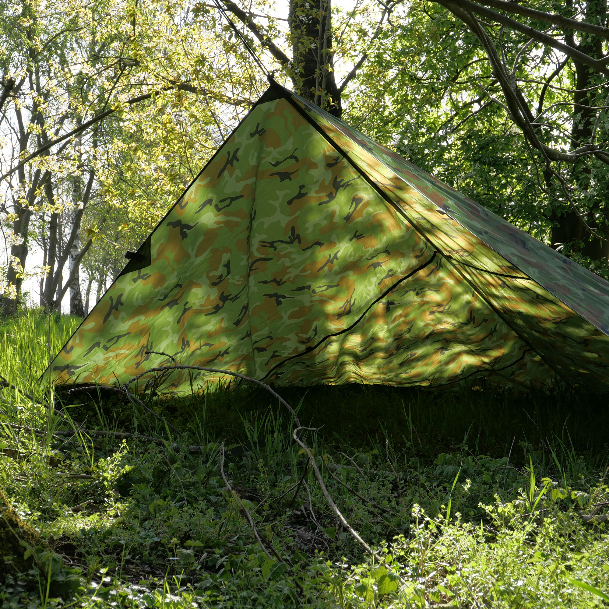 Telo yourGEAR Bushcraft Tarp 3x3 mimetico montato tra alberi, offre protezione impermeabile e UV 50+ per campeggio, trekking o survival. Struttura robusta, ideale come tenda, tettoia o riparo all'aperto.