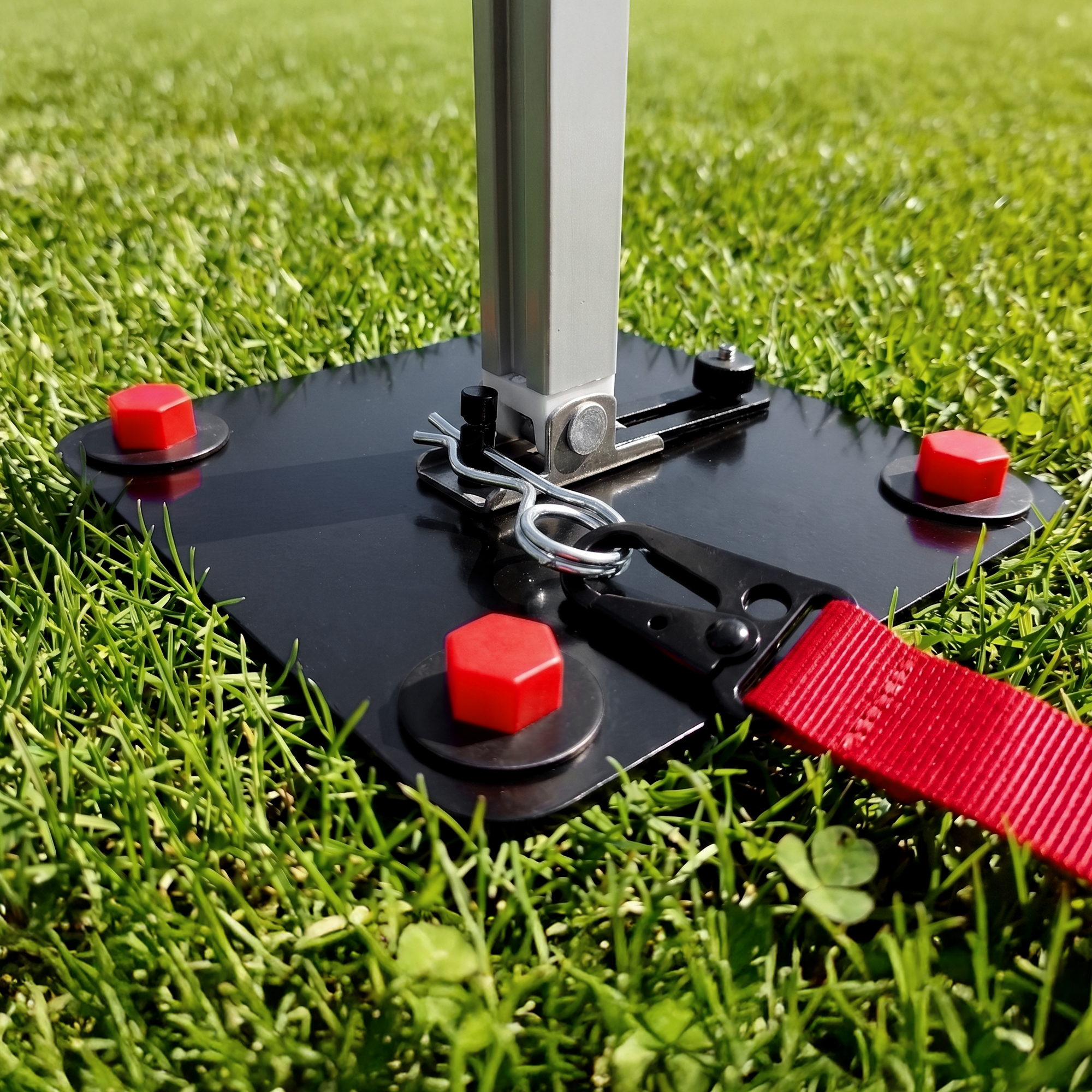 Close-up of your GEAR Montana black anchor plate on grass with attached silver telescopic awning foot, red plastic locking caps, metal quick-release pins and red strap for secure caravan/awning fixation.