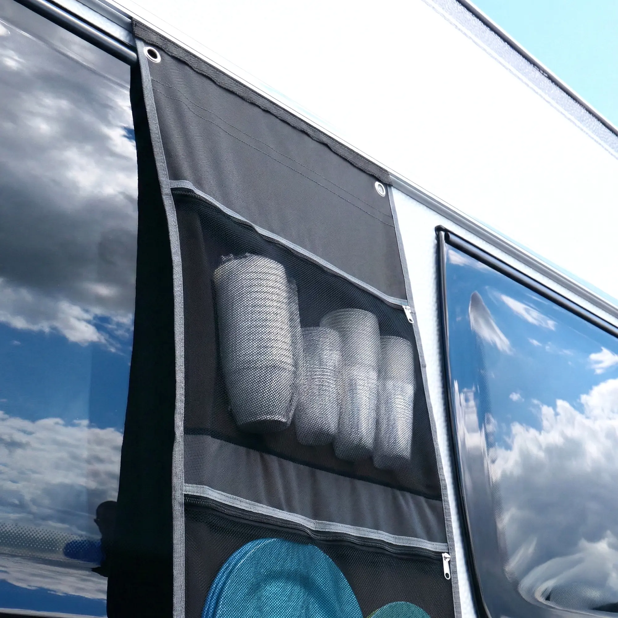 Organiseur your GEAR en tissu noir à quatre poches filet suspendu sur la paroi d’un camping‑car, œillets et fermeture éclair visibles, poche supérieure contenant gobelets cylindriques et poche inférieure avec assiettes bleues.