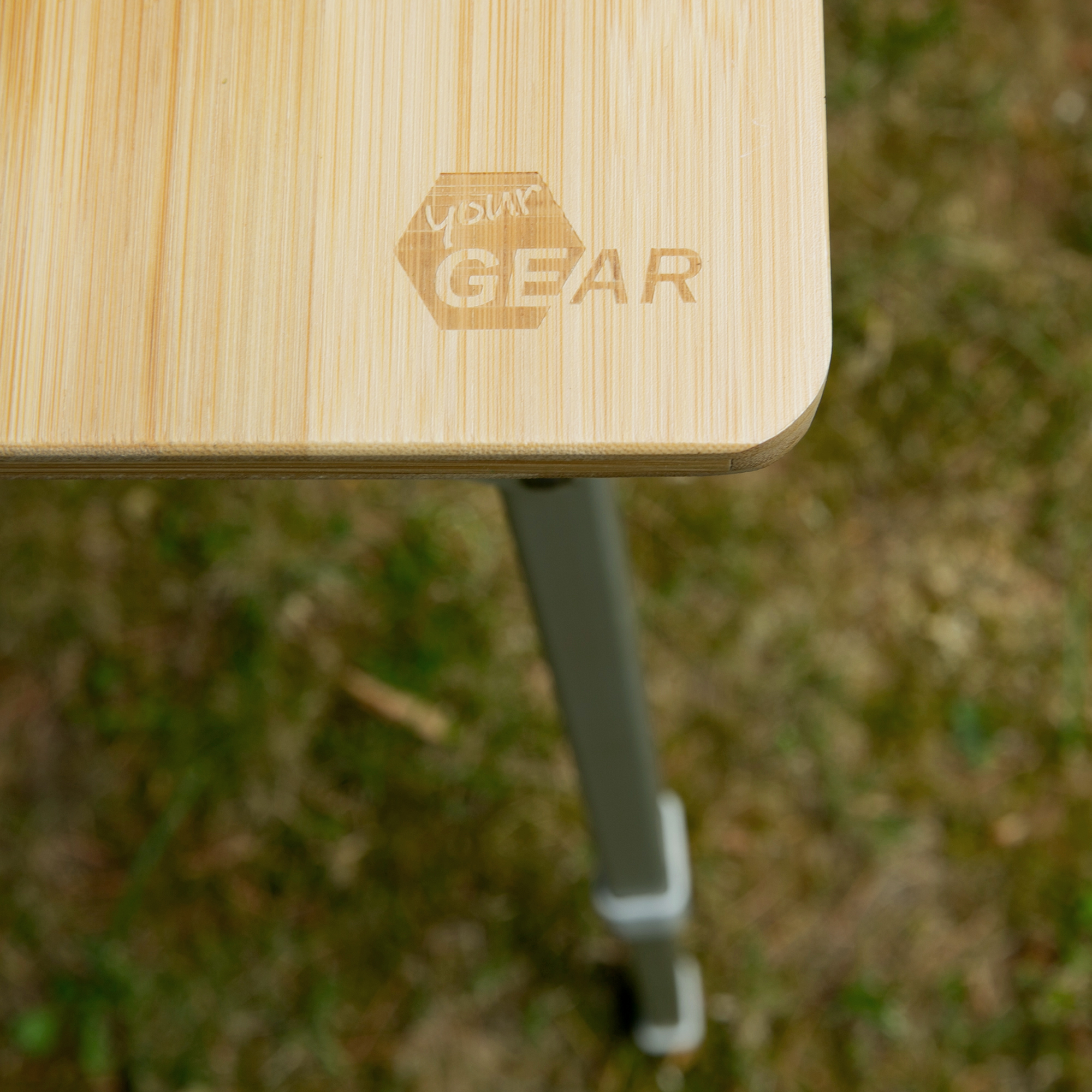 Close-up of your GEAR Portofino XL bamboo camping table, showing the natural bamboo tabletop with engraved logo and a sturdy, height-adjustable metal leg, suitable for outdoor use by up to 6 people.