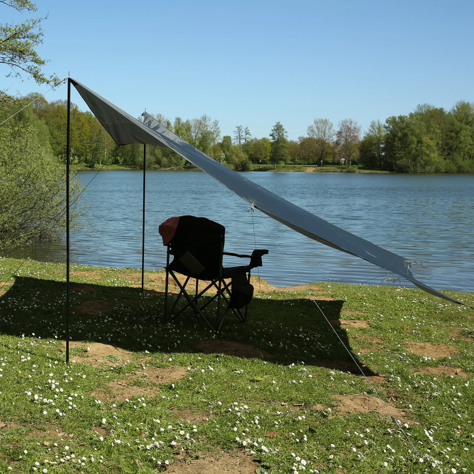 Gray 3x3m your GEAR Atrani sunshade tarp set up by a lakeside with two black support poles, guy lines and a folding chair underneath, waterproof fabric and UV50+ protection visible in outdoor camping scene