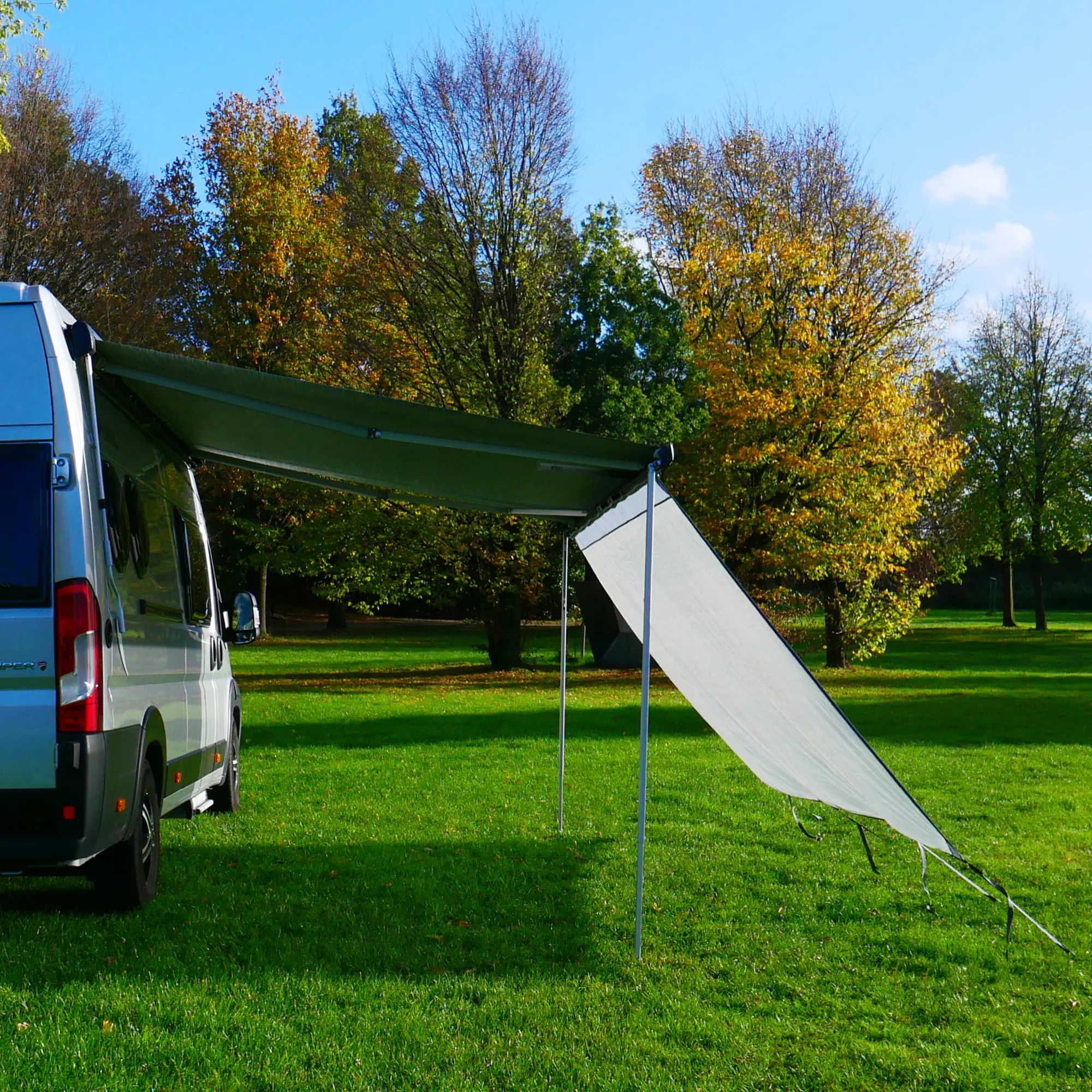 Wohnmobil mit ausgefahrener Markise und your GEAR Capri Sun Blocker 2,8 x 2 m als Vorderwand; lichtdurchlässiges Gewebe bietet Sonnen- und Sichtschutz, befestigt an der Markise, auf einer Wiese im Freien.