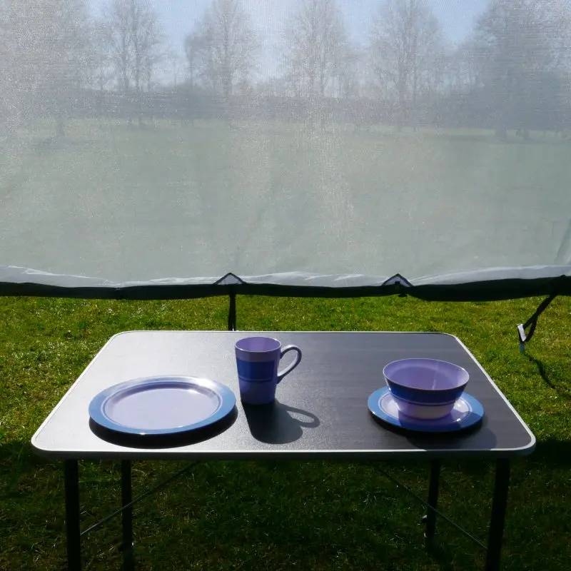 Vorderwand your GEAR Sun + Rain Blocker 3,7 x 2 m an Markise montiert, lichtdurchlässiges Spezialgewebe, schützt vor Sonne und Regen; davor Campingtisch mit blauem Geschirr auf Wiese.