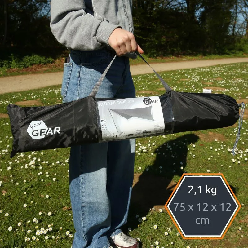 Sac de transport noir porté à la main contenant un Sonnensegel your GEAR "Atrani 3x3" avec étiquette visible, sangle grise, poids 2,1 kg indiqué, tissu gris-beige, idéal pour camping/soleil, photo prise en plein air sur pelouse.