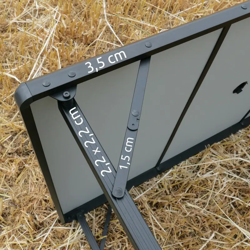 Dessous du your GEAR Campingtisch Tivoli XL : structure en aluminium noir avec mesures 3,5 cm, 2,2 x 2,2 cm et 1,5 cm, plateau gris, pieds pliables, posé sur de l’herbe sèche. Table de camping imperméable.