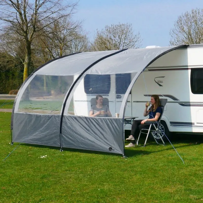 Toldo/Vorzelt gris claro your GEAR Riviera 200x260 cm unido a caravana, estructura arqueada con dos arcos, paneles laterales transparentes grandes, protección UV, suelo abierto, personas sentadas debajo con mesa y sillas.