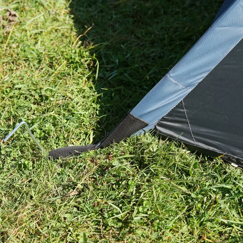 Détail du your GEAR Zelt Monza pour 4 personnes : coin de la toile extérieure bleue et noire fixé au sol avec une sangle et un piquet, montrant la solidité et la facilité de montage du camping.