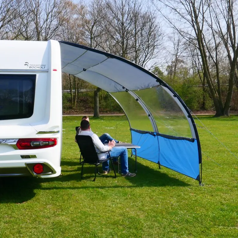 Vorzelt your GEAR Riviera 300 am Wohnwagen, 3 x 2,6 m, mit UV 50+ Sonnenschutz, Panoramafenster und blauem Windschutz. Zwei Personen sitzen darunter auf Campingstühlen an einem Tisch.