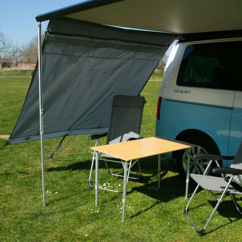 Side wall for camper van awning, your GEAR Sun + Rain Blocker, grey double-layered fabric, mounted on awning arm, providing wind and rain protection; camping chairs and table set up underneath.