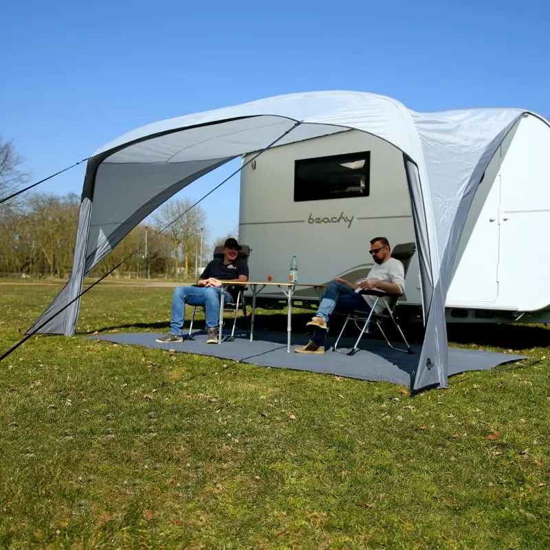 Auvent your GEAR Amalfi Air 360 gris, gonflable, monté sur une caravane Beachy, offrant une grande zone d’ombre. Deux personnes sont assises sous l’auvent avec table et chaises sur une pelouse.