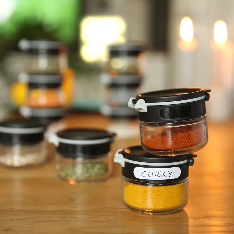 your GEAR Double-Flip spice jars set, 12 glass containers (6 x 200 ml, 6 x 85 ml) with black lids, stackable, labeled, shown on a wooden kitchen table for organized spice storage.