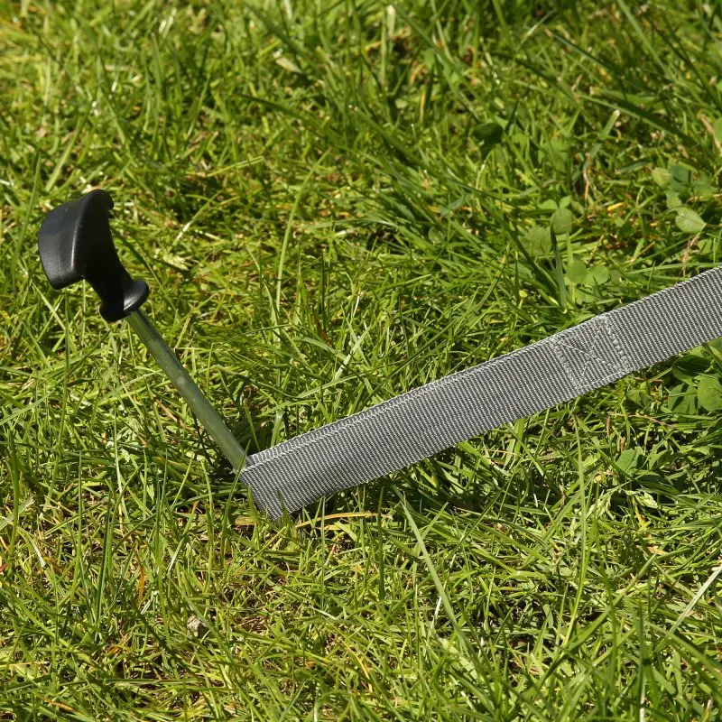 Sangle tempête grise your GEAR de 25 mm fixée au sol avec un piquet à poignée noire, idéale pour tentes, auvents et accessoires camping, posée sur de l’herbe verte.