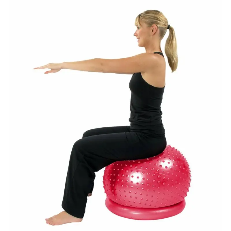 Woman sitting on a red Carnegie massage ball with 65 cm diameter and nubs, placed on a stabilizing ring; suitable for fitness, self-massage, and gymnastics. The ball is shown with textured surface and included base.