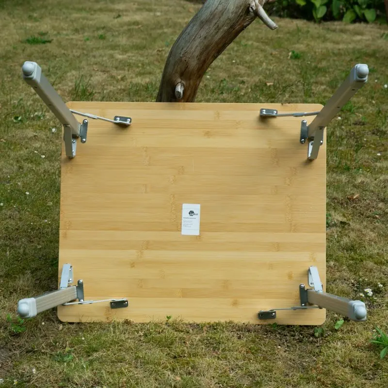 Bambus Campingtisch your GEAR Portofino M mit klappbaren, höhenverstellbaren Aluminiumbeinen, robuste 1,2 cm Bambus-Tischplatte, geeignet für 2 Personen, auf einer Wiese stehend, Unterseite sichtbar.