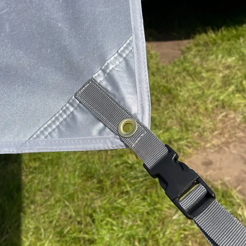 Corner of your GEAR Caravan Thermo Tarp with reflective aluminum-coated Oxford polyester, metal eyelet, and adjustable strap with black plastic buckle, shown outdoors on grass. UV80, waterproof, sun protection awning.