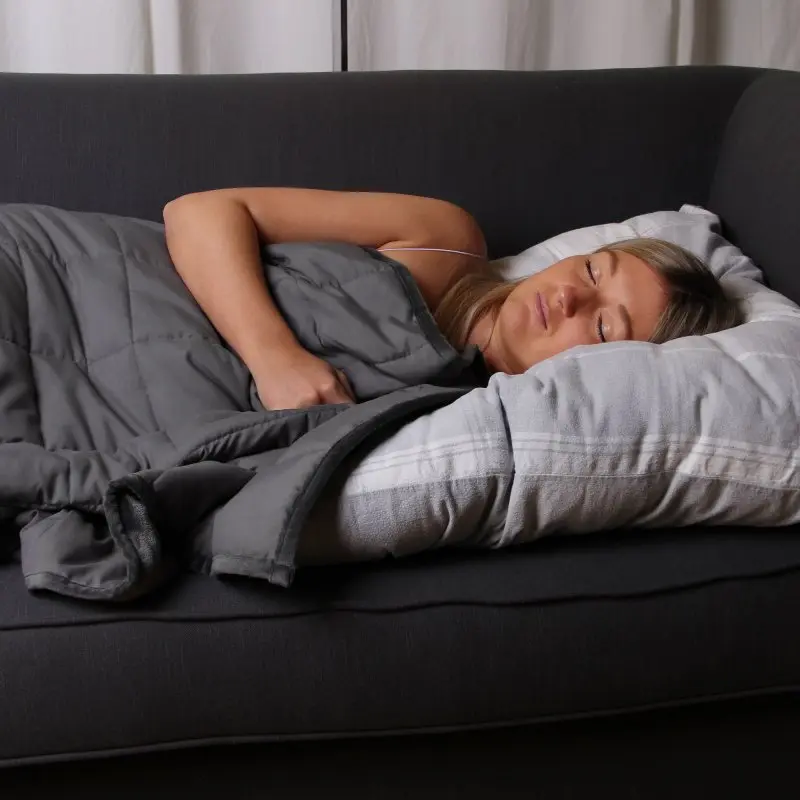 Mujer descansando en un sofá bajo una manta terapéutica yourGEAR Cameri de 6 kg, color gris, tamaño 135x200 cm, de terciopelo, diseñada para aliviar el estrés y mejorar el sueño mediante presión profunda.