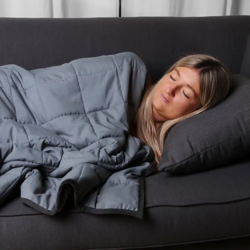 Woman resting on a sofa covered with the yourGEAR Como 9 kg weighted therapy blanket, 135x200 cm, made of grey quilted cotton, designed for deep pressure stimulation and relaxation.