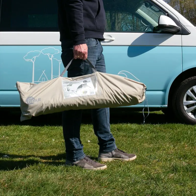 Persona che tiene la borsa da trasporto del your GEAR Tropea TC Bus Vorzelt, un tarp parasole in tessuto misto cotone UV 50+ per van, davanti a un camper parcheggiato su prato verde.