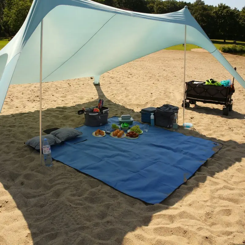 Large yourGEAR Blue XL picnic blanket, 200x200cm, laid out on sand under a sunshade, with food, pillows, and picnic gear on top; soft fleece top, waterproof and insulated underside visible.
