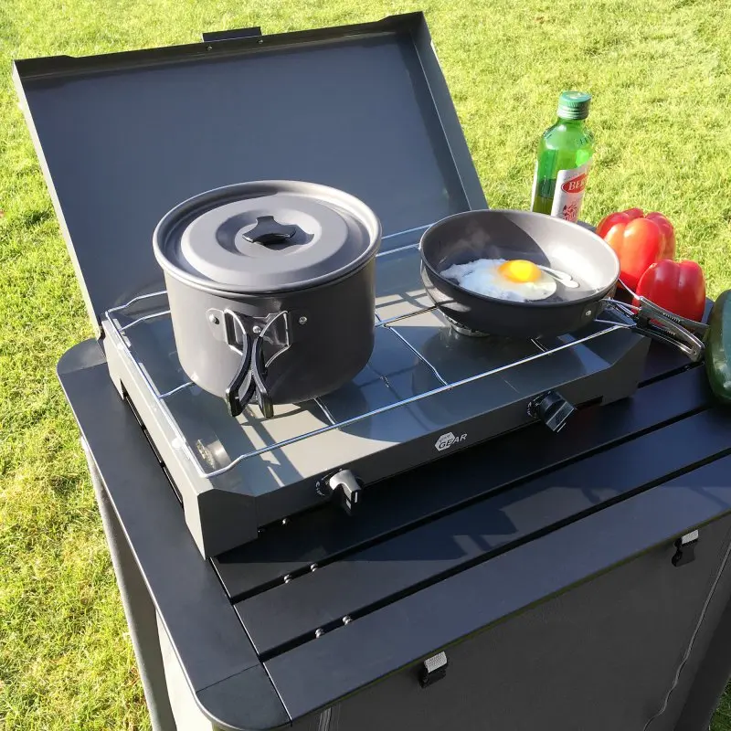 Set de cuisson camping your GEAR Ranger V pour 4-5 personnes, avec grande casserole à couvercle et poêle sur réchaud, poignées pliables, légumes et bouteille d’huile à côté, idéal pour l’extérieur.