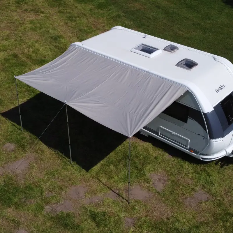 Wohnwagen mit your GEAR Caravan Tarp TC 450 x 240 Sonnendach aus Baumwollmischgewebe, befestigt am Caravan, drei Alu-Teleskopstangen sorgen für Stabilität und großzügigen Schatten auf einer Wiese.
