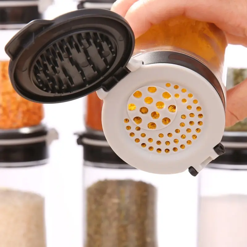 Glass spice jar from your GEAR Double-Flip set, open to show dual flip lid with shaker and pour options, filled with yellow spice. Other spice jars are visible in the background. Ideal for kitchen organization.