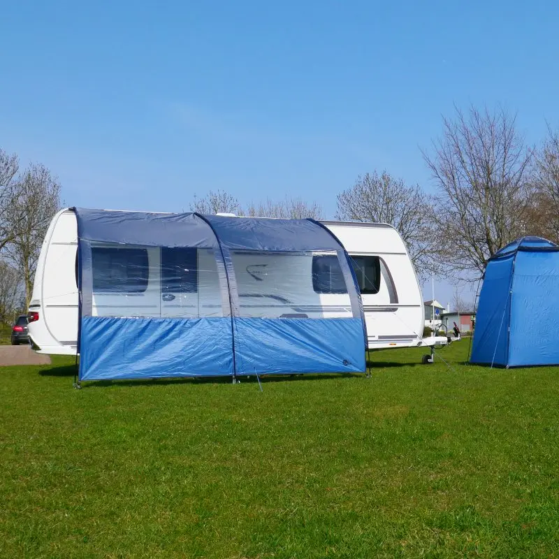 your GEAR Riviera 300 Vorzelt, 3 x 2,6 m, als Vordach am Wohnwagen montiert, mit großen Panoramafenstern, UV 50+ Sonnenschutz, blau-graues Wetterschutzdach auf grüner Wiese bei Sonnenschein.