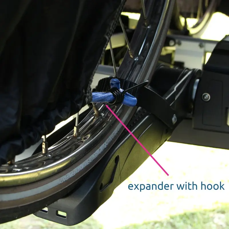 Close-up of yourGEAR Malaga bike cover secured on a rear bike rack, showing waterproof fabric, wheel, and expander with hook fastening the cover to the bicycle for protection during transport.