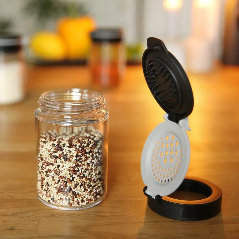your GEAR Double-Flip spice jar made of clear glass, open black and white flip lid with shaker holes, filled with mixed spices, shown on a wooden kitchen surface with blurred jars in the background.