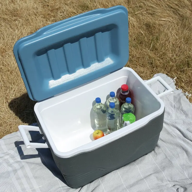 Caja térmica your GEAR Yukon de 66 L abierta sobre una manta, mostrando varias botellas, una manzana y un diseño robusto con aislamiento grueso, tapa azul y asas blancas, ideal para mantener bebidas frías.
